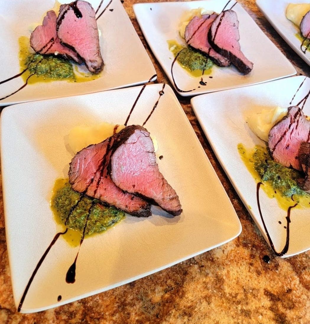 Square white plates lined up on a stone counter, each with slices of rare seared beef tenderloin over a green herb sauce with a balsamic drizzle.