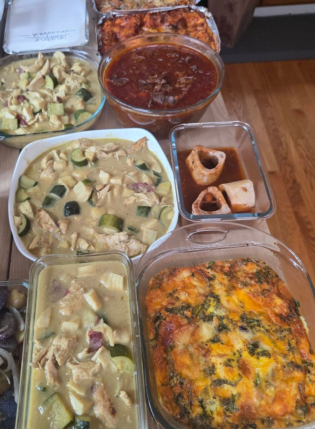 Meals laid out on a wooden table: trays of lasagna, dishes of green chicken curry with zucchini, a bowl of red stew, a small dish of marrow bones, a frittata and roasted potatoes.